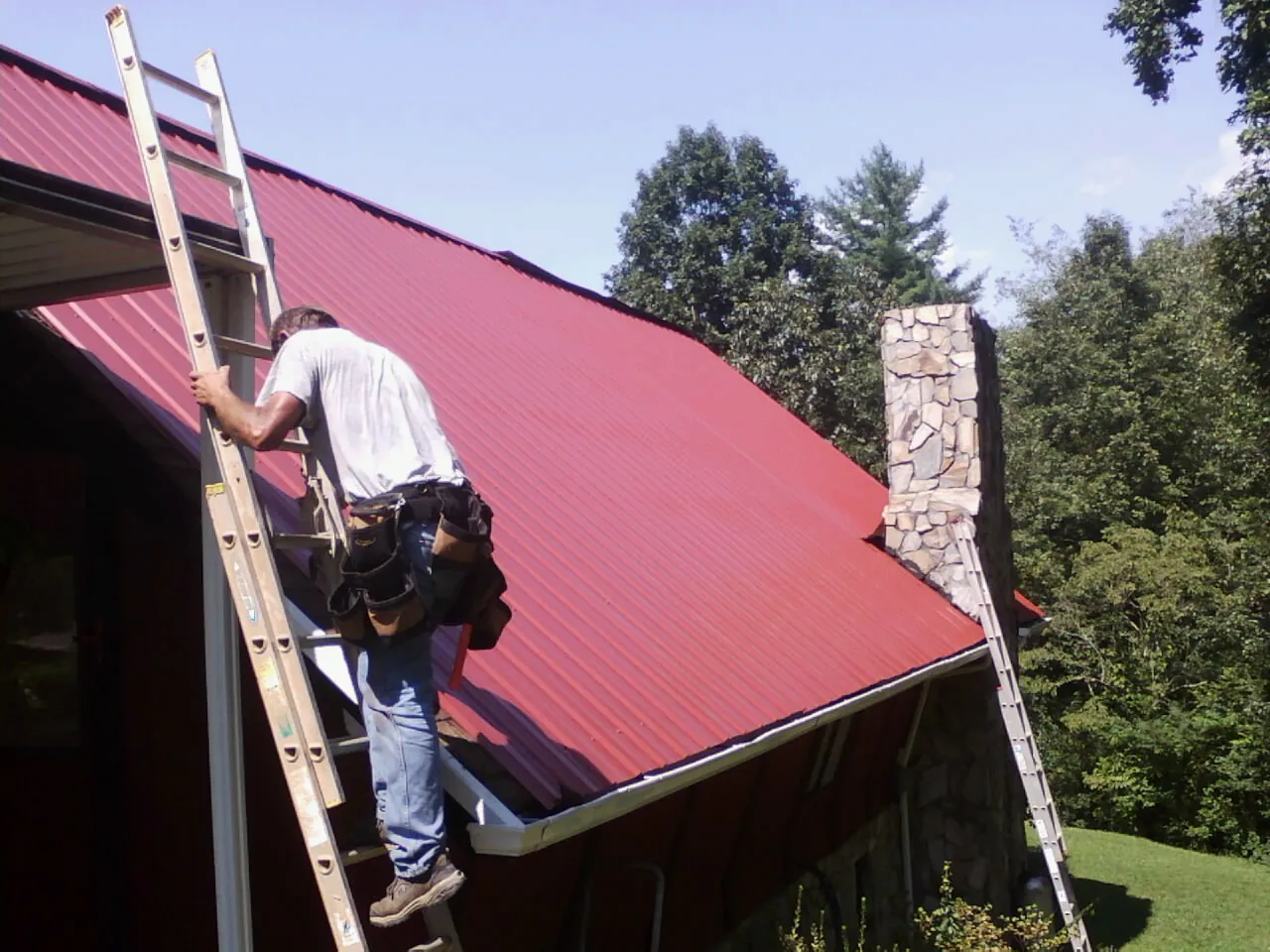 Roofer on a ladder beside a red metal roof and stone chimney — Soffit & Fascia Repair by Roofing Brothers in Parkesburg